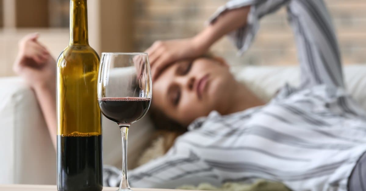A close-up of a bottle of wine next to a half-poured glass on the coffee table. A woman lays on the couch behind them