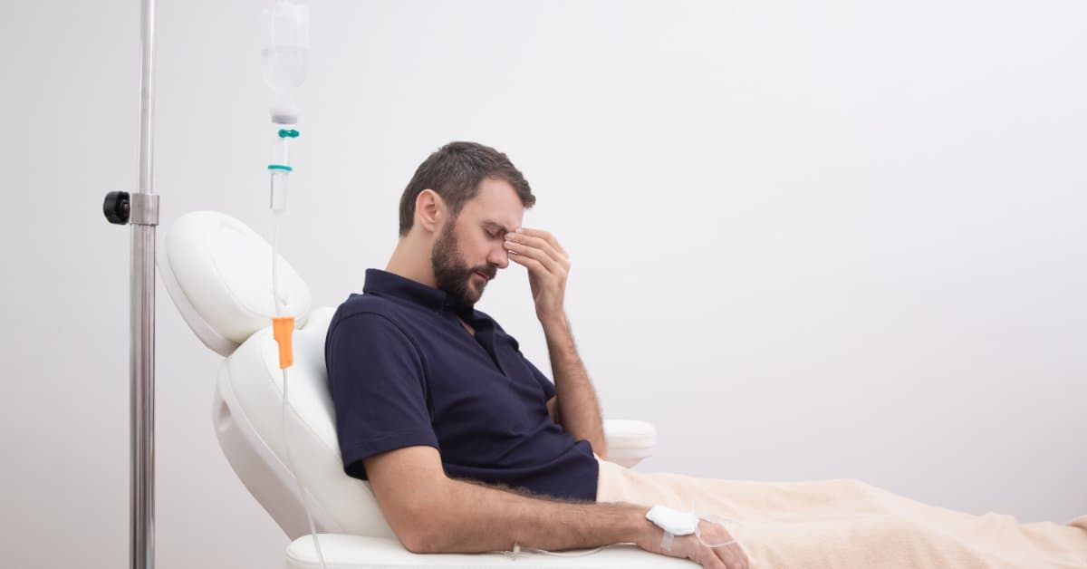 A man with a hangover sits in a recliner with his head in his hand. He receives an IV hydration therapy treatment.
