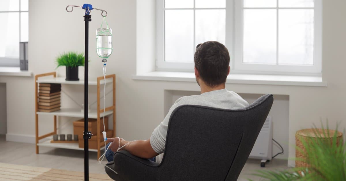 A man sits in his living room as he faces the windows. He receives an IV drip treatment, sitting in an accent chair.