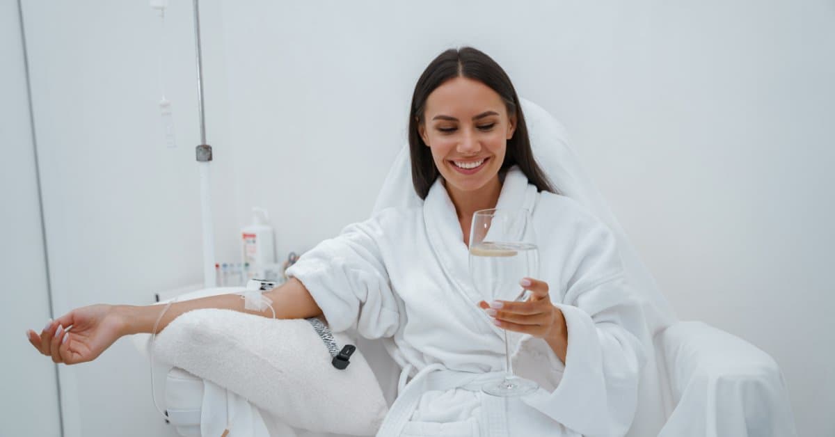 A young woman sits in a chair and smiles at her phone in a white robe as she receives a mobile IV drip.
