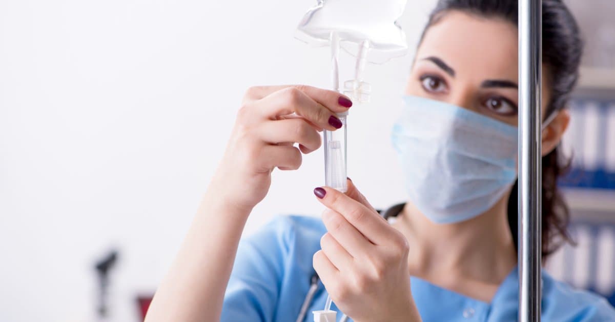 A nurse wears a face mask and blue scrubs as she places an IV therapy drip for an in-home IV therapy session.