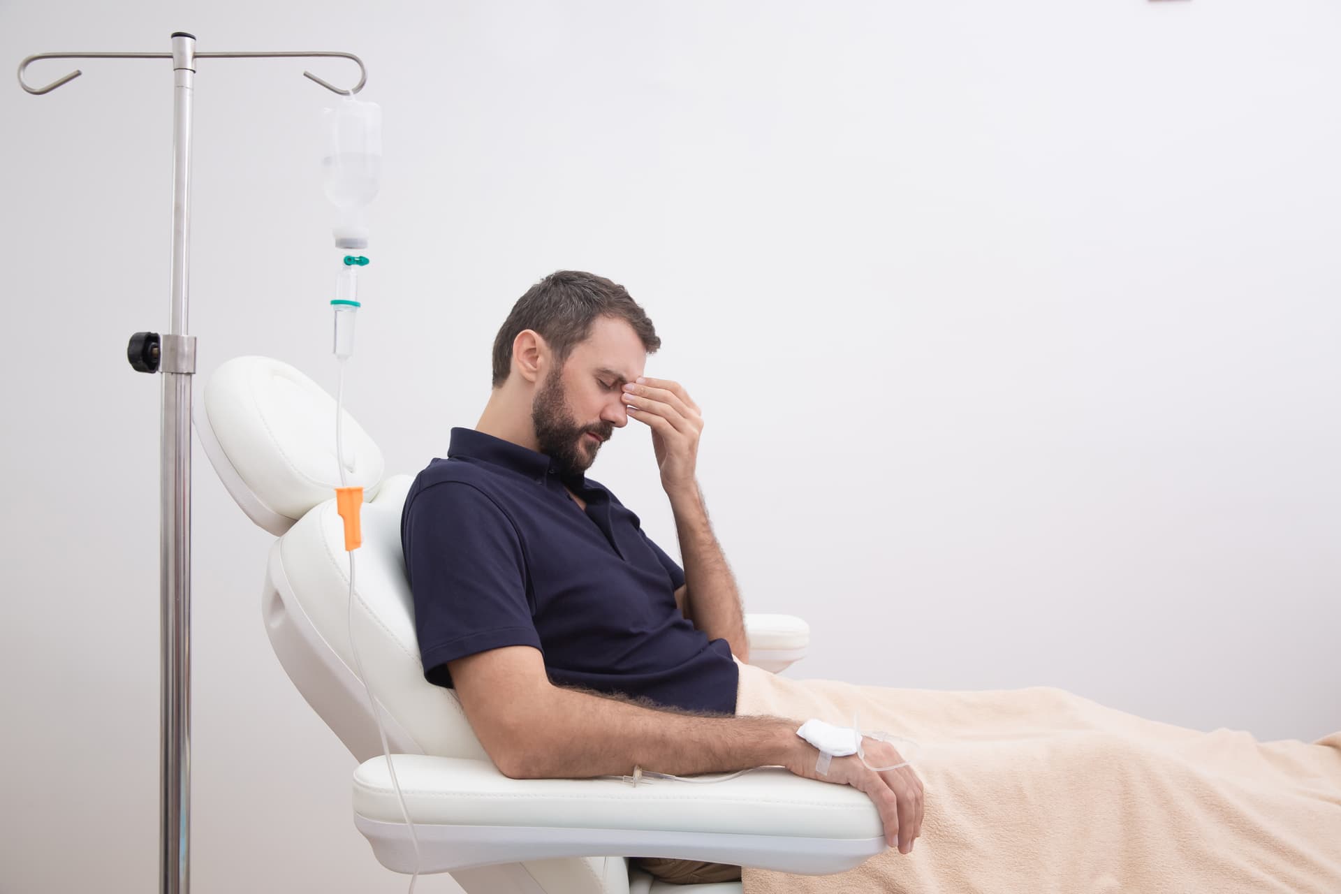 An adult male sits in an accent chair with his head in his hand as he receives an IV trip for his hangover.