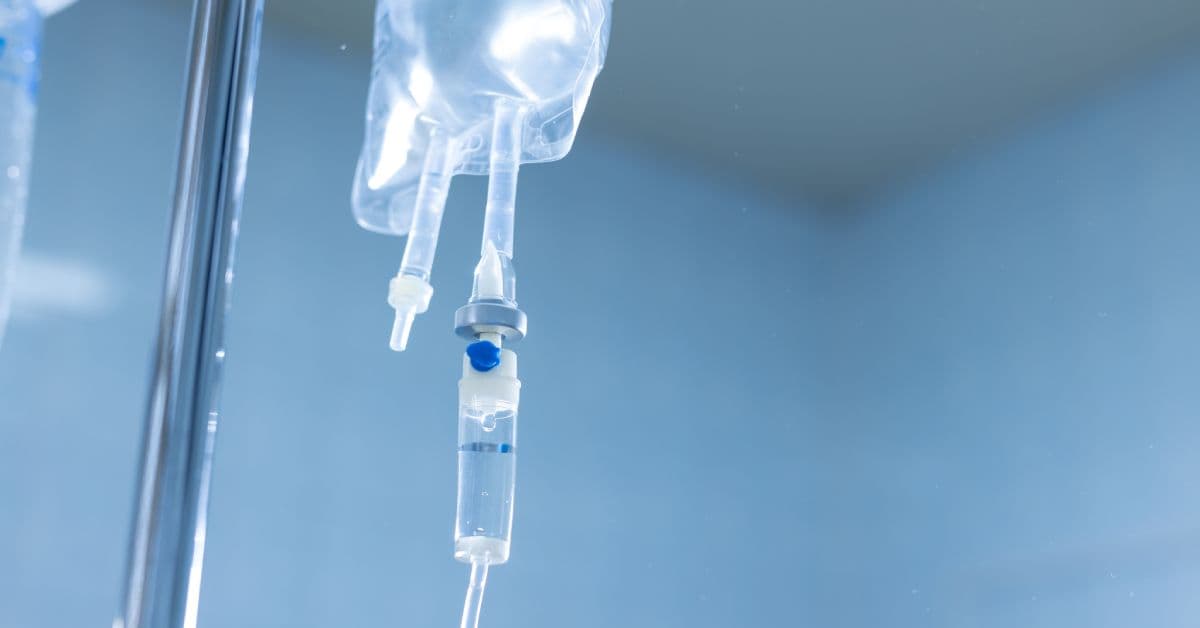 A close-up of an IV drip setup with a silver pole and a clear fluid in the bag. The IV bag is also clear.