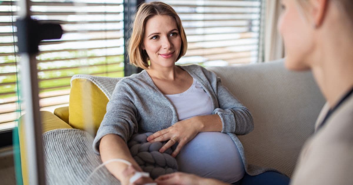 A pregnant woman comfortable at home in a sitting chair holding her belly as someone administers an IV hydration drip.