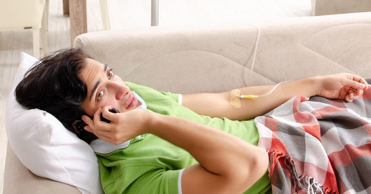 A young man at home on the couch talks on the phone as he receives a mobile IV drip. He's in a plaid blanket.