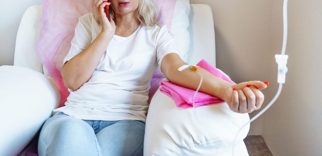 A woman sits in a comfortable chair as she receives an IV drip therapy treatment at home. She talks on the phone.