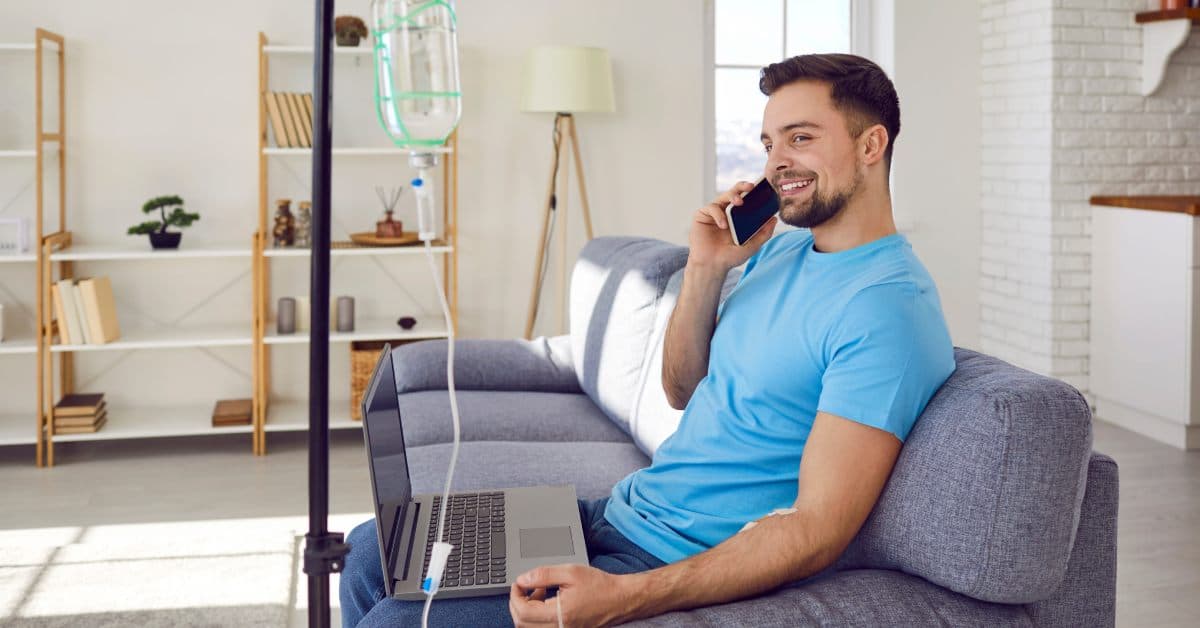 A young man sits on his couch in a blue T-shirt as he talks on the phone and receives mobile IV therapy.