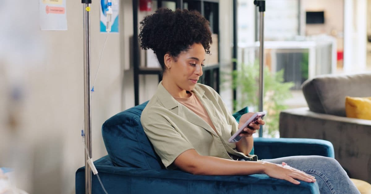 A Black woman sits in an accent chair as she looks at her phone and receives a mobile IV drip in her home.