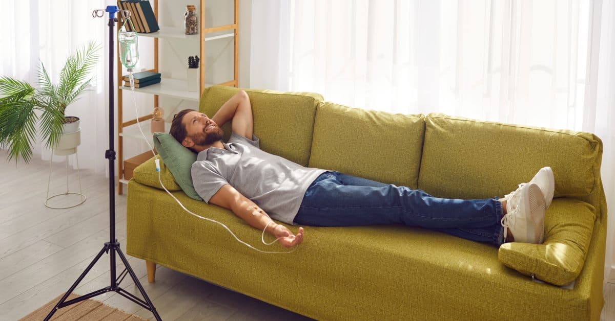 A man lays on a green couch as he receives an IV drip in his home. His left arm is folded behind his head.
