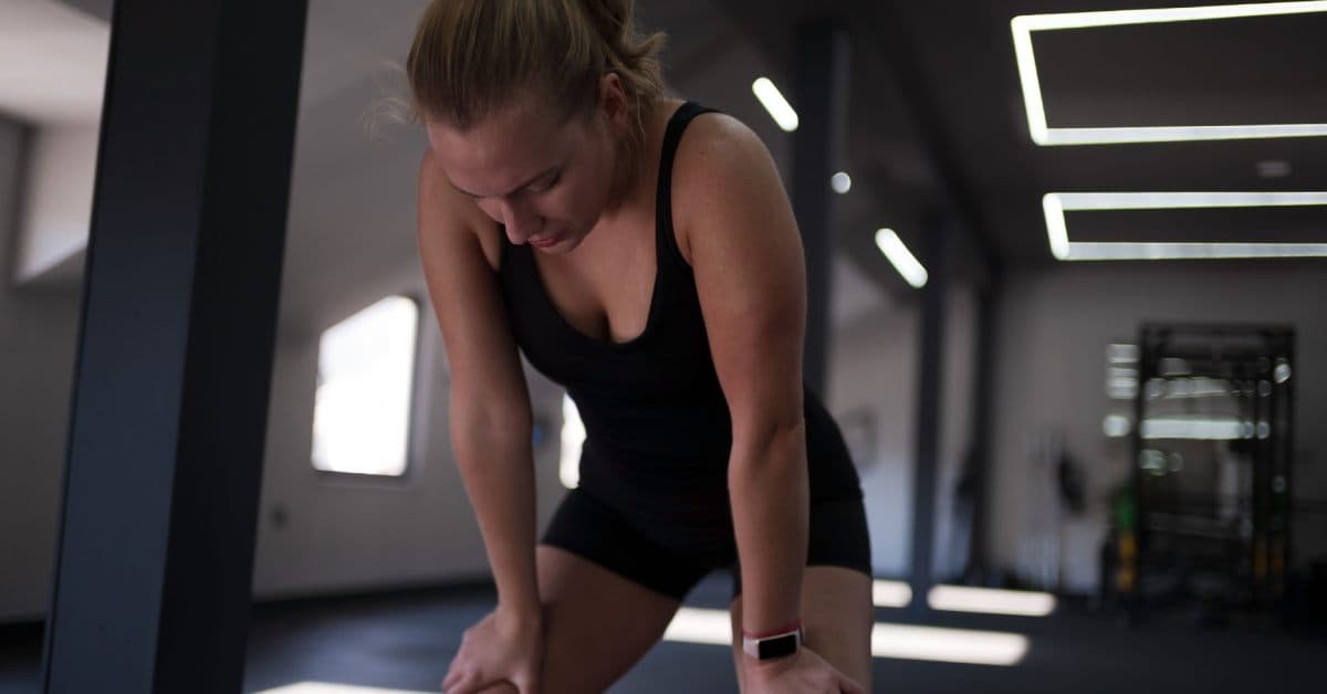 A female athletic kneeling in the gym with her hands on her knees. She's catching her breath after a workout.