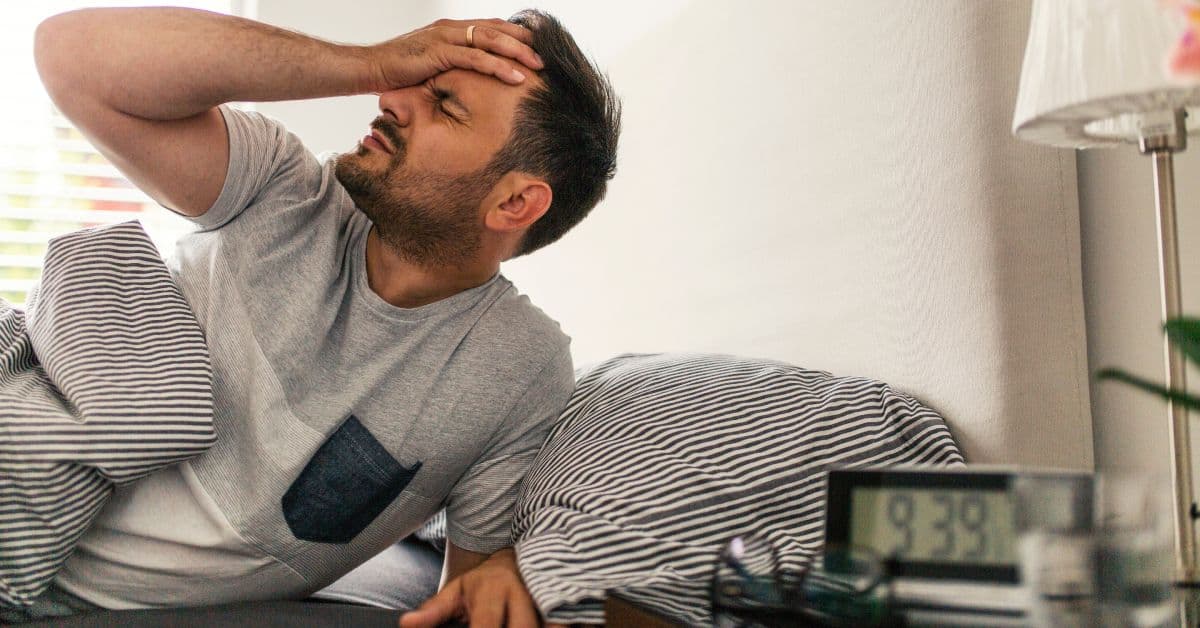 A man waking up in his bed with a headache. He's holding his head with one hand and water sits next to the alarm clock.