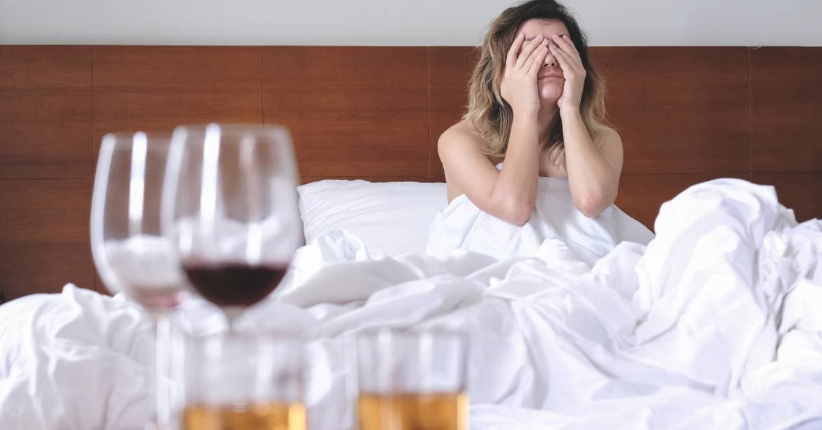 A woman wakes up in bed with her hands over her face to rub her eyes. Two wine glasses and whiskey glasses sit on the table.