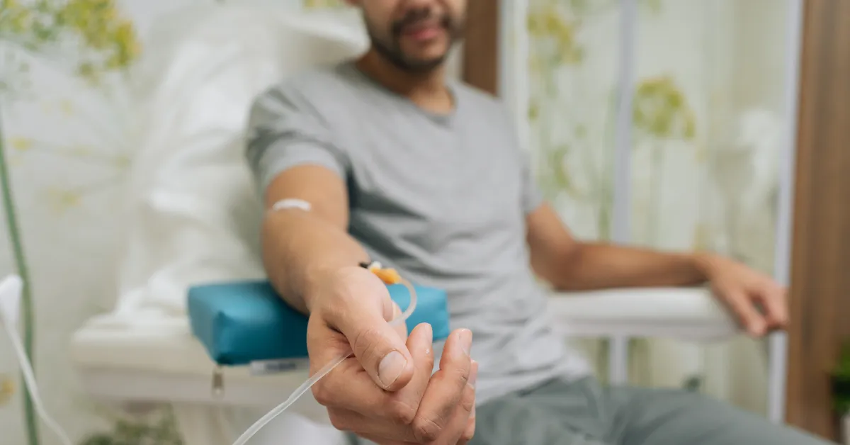 A patient sits in a clinic chair as he receives IV therapy through his veins. He sits in casual clothes.