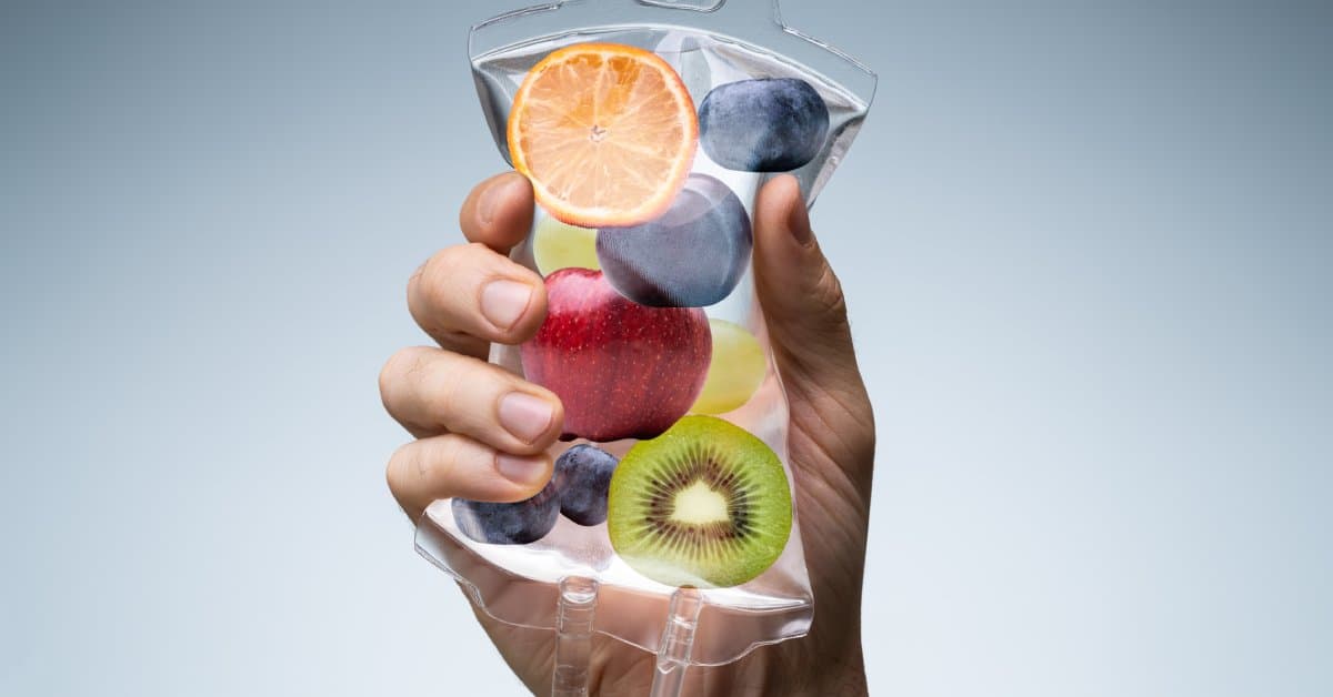 A close-up of a hand holding a bag of IV fluid with fruit inside the fluid. The fruit symbolizes vitamin content in the fluid.