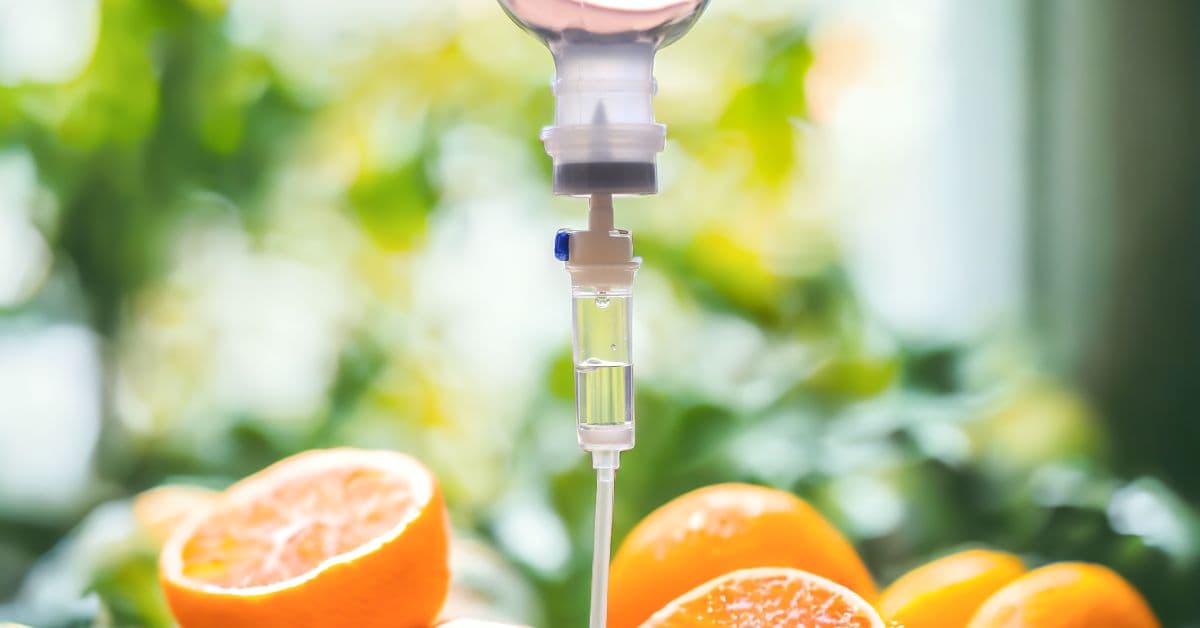 A pile of oranges sits underneath an IV drip setup. The drip is administering vitamins and minerals.