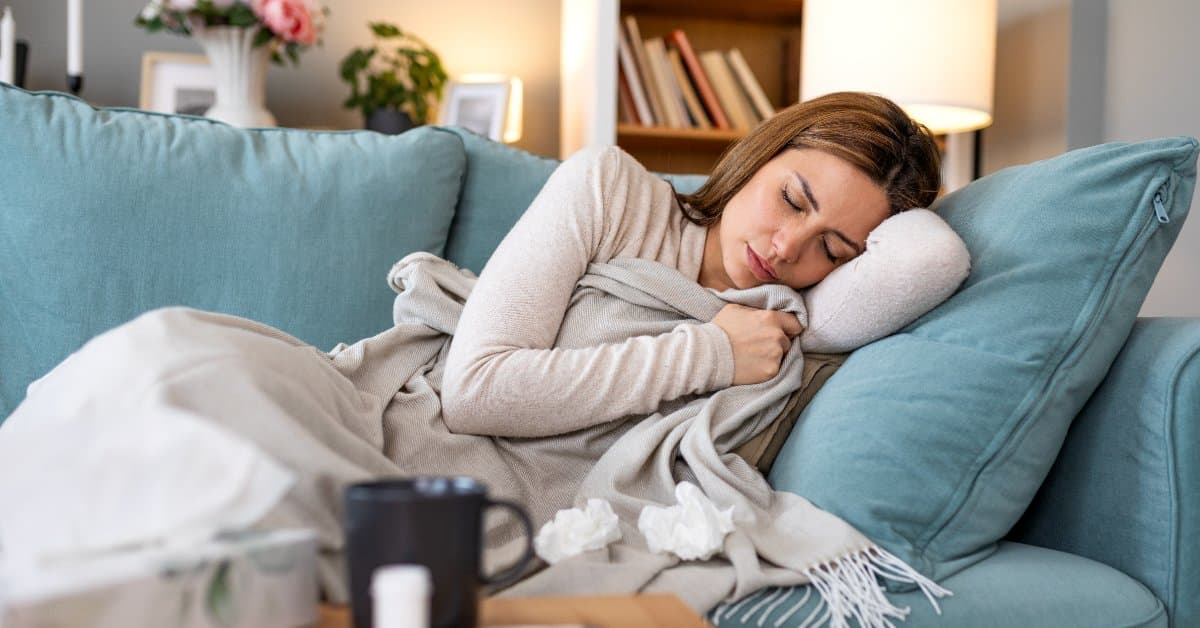 A sick woman lays at home on the couch under a blanket as she takes a nap. A thermometer and a coffee mug sit on the table.