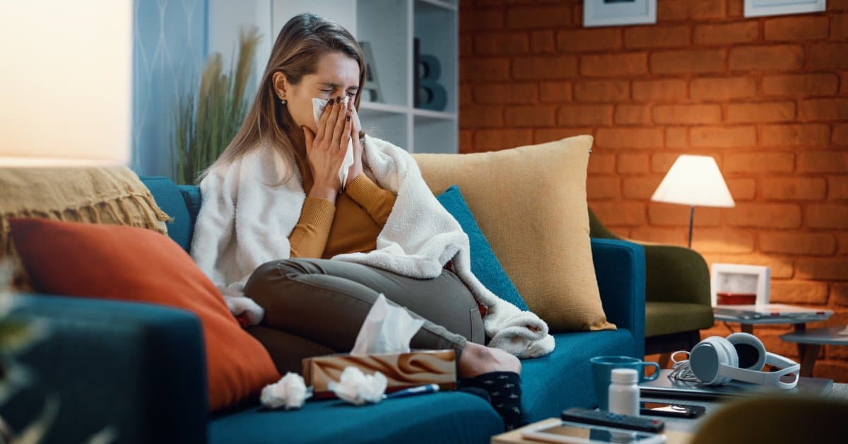 A young woman at home on her blue couch curled up in a blanket as she blows a runny nose into a tissue.