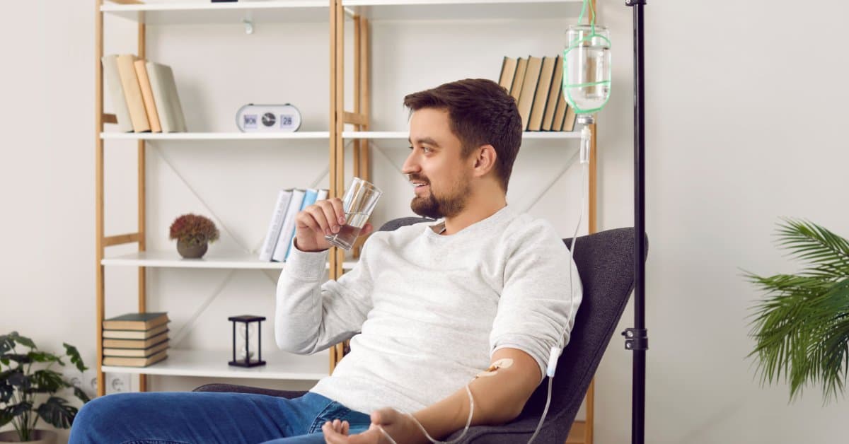 A man in the comfort of his own home receives an IV therapy treatment in a recliner chair. He drinks water.
