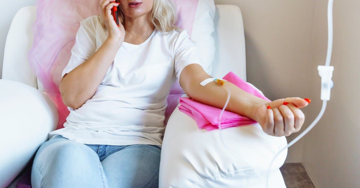 A close-up of a woman in a comfortable chair at home receives an IV treatment as she talks on the phone